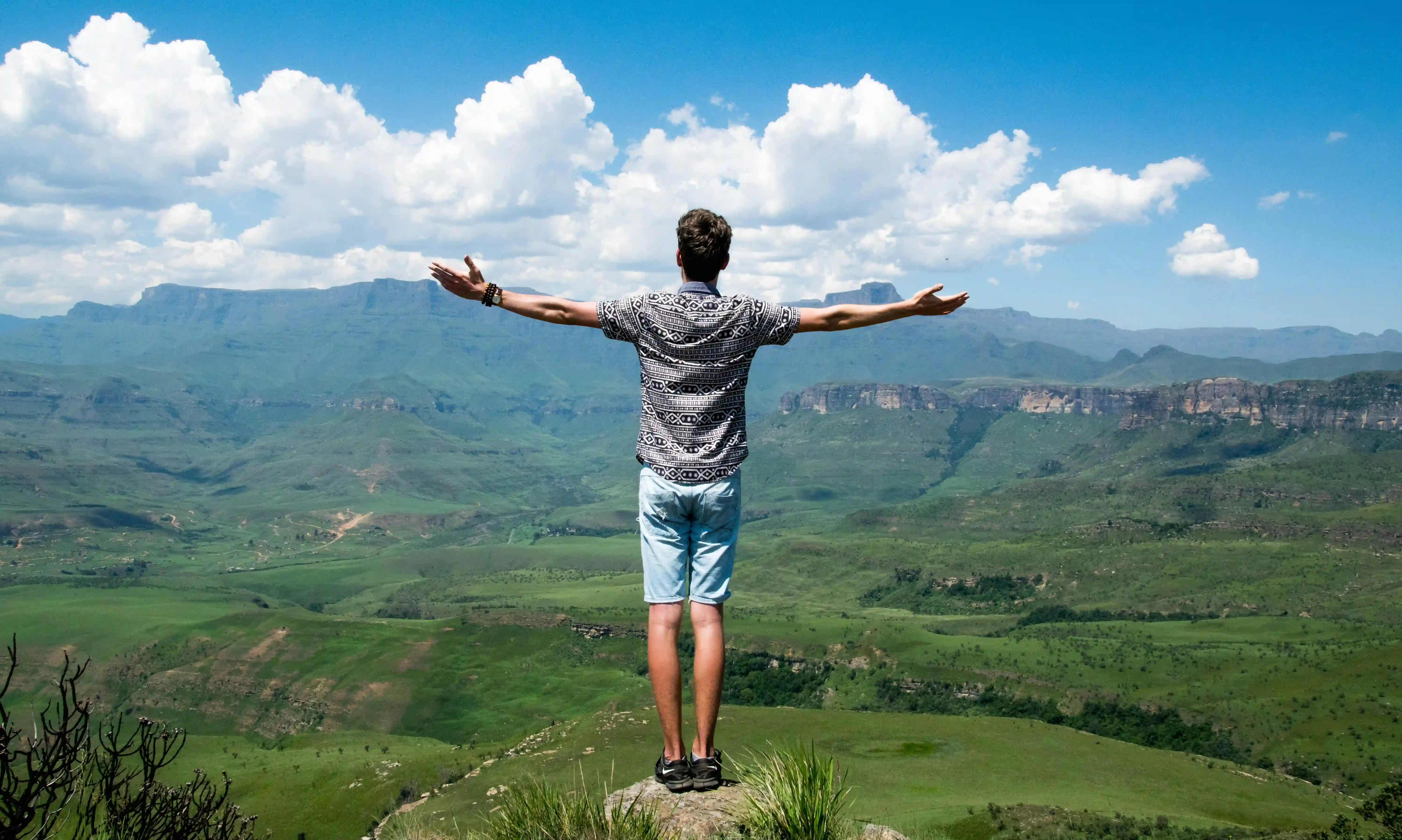 Person standing on a rock with arms outstretched facing a vast green mountainous landscape under a blue sky with scattered clouds.