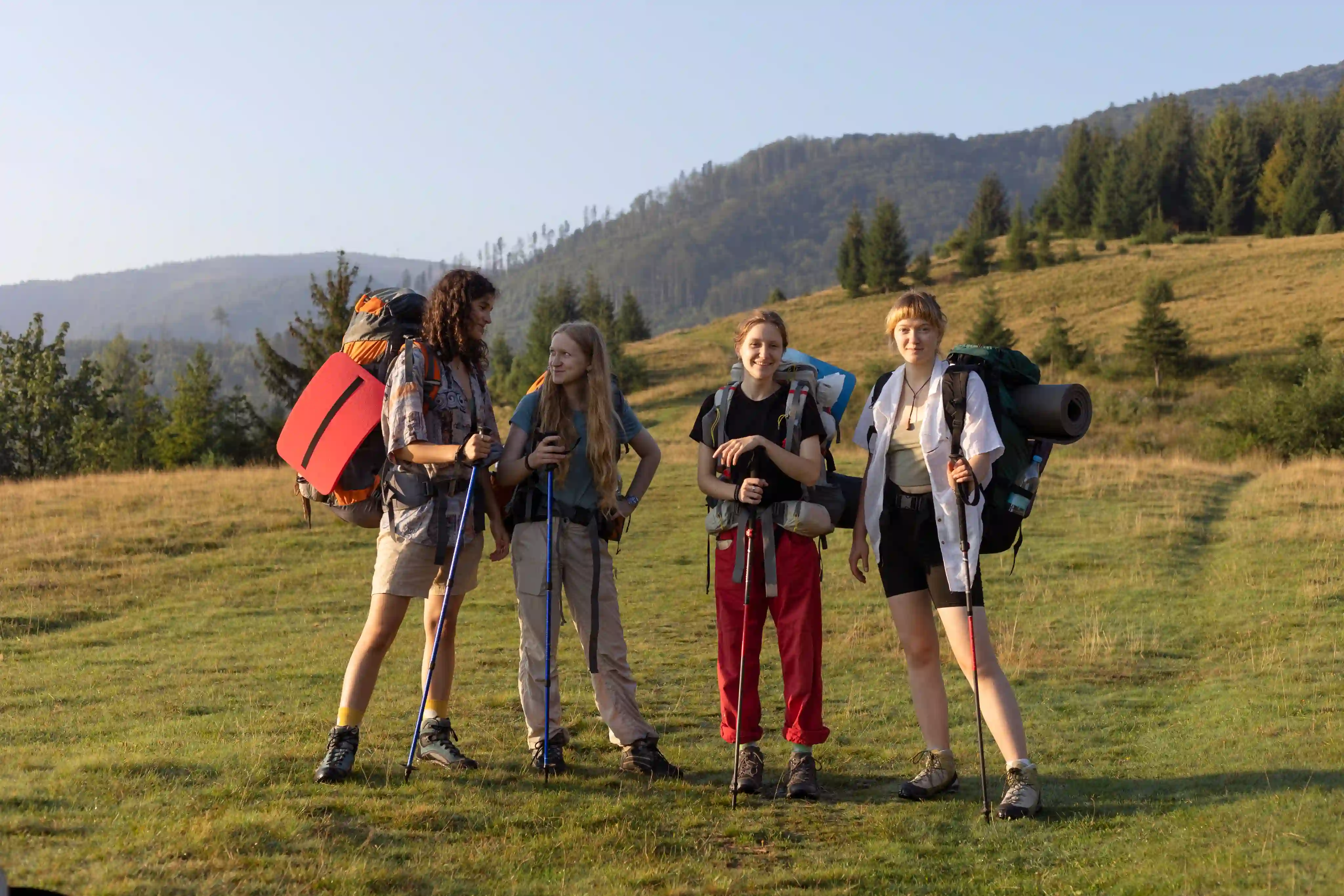 Four young hikers with backpacks and trekking poles standing on a grassy field with forested hills in the background.