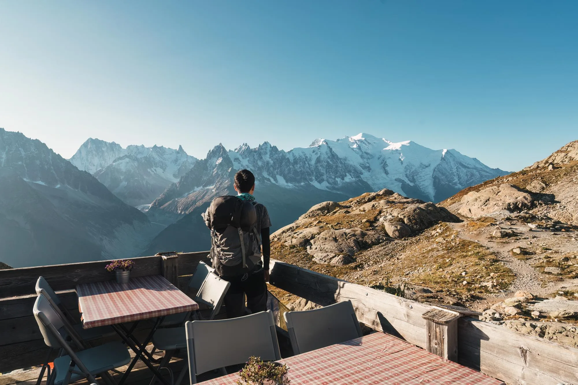 Hiker with backpack standing on a terrace overlooking snow-covered mountain peaks under a clear blue sky.
