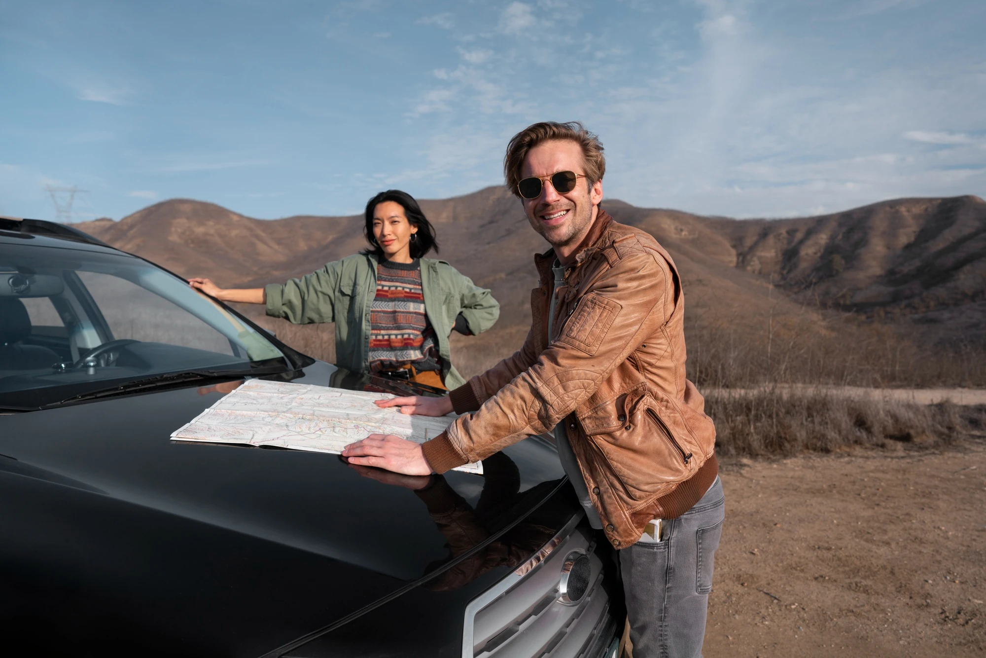 Man in a brown jacket and sunglasses leaning on a black car with a map spread on the hood, woman standing beside the car with hand on hip in a desert landscape with mountains.