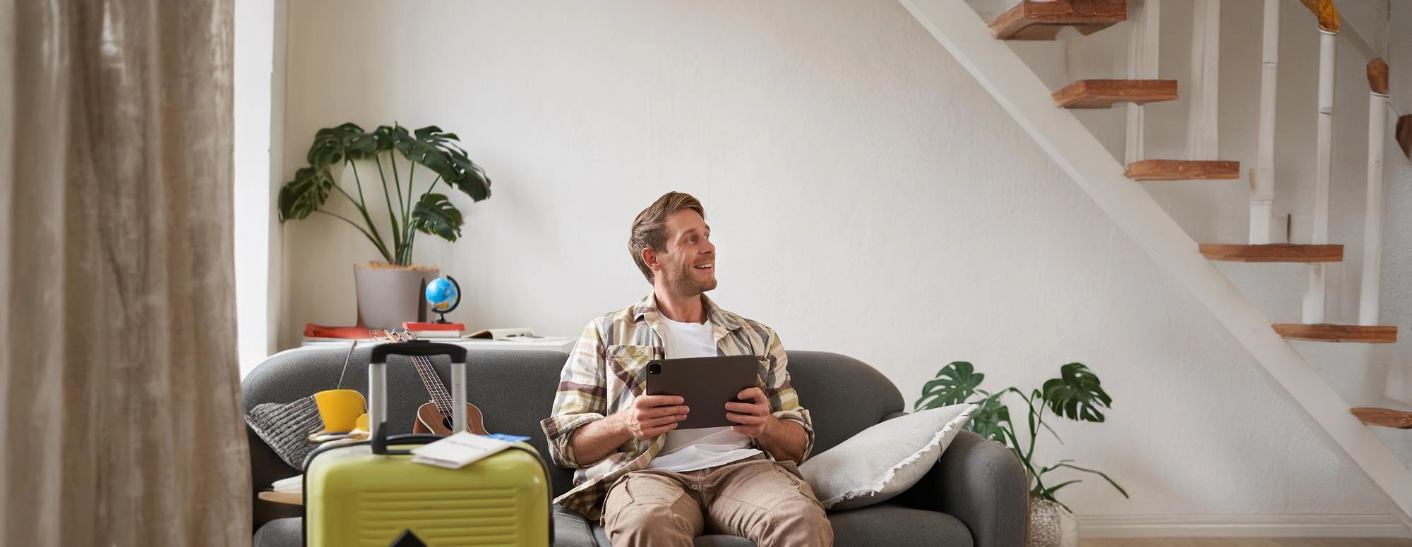 Man sitting on gray couch holding a tablet with an open yellow suitcase and guitar nearby in a cozy living room.