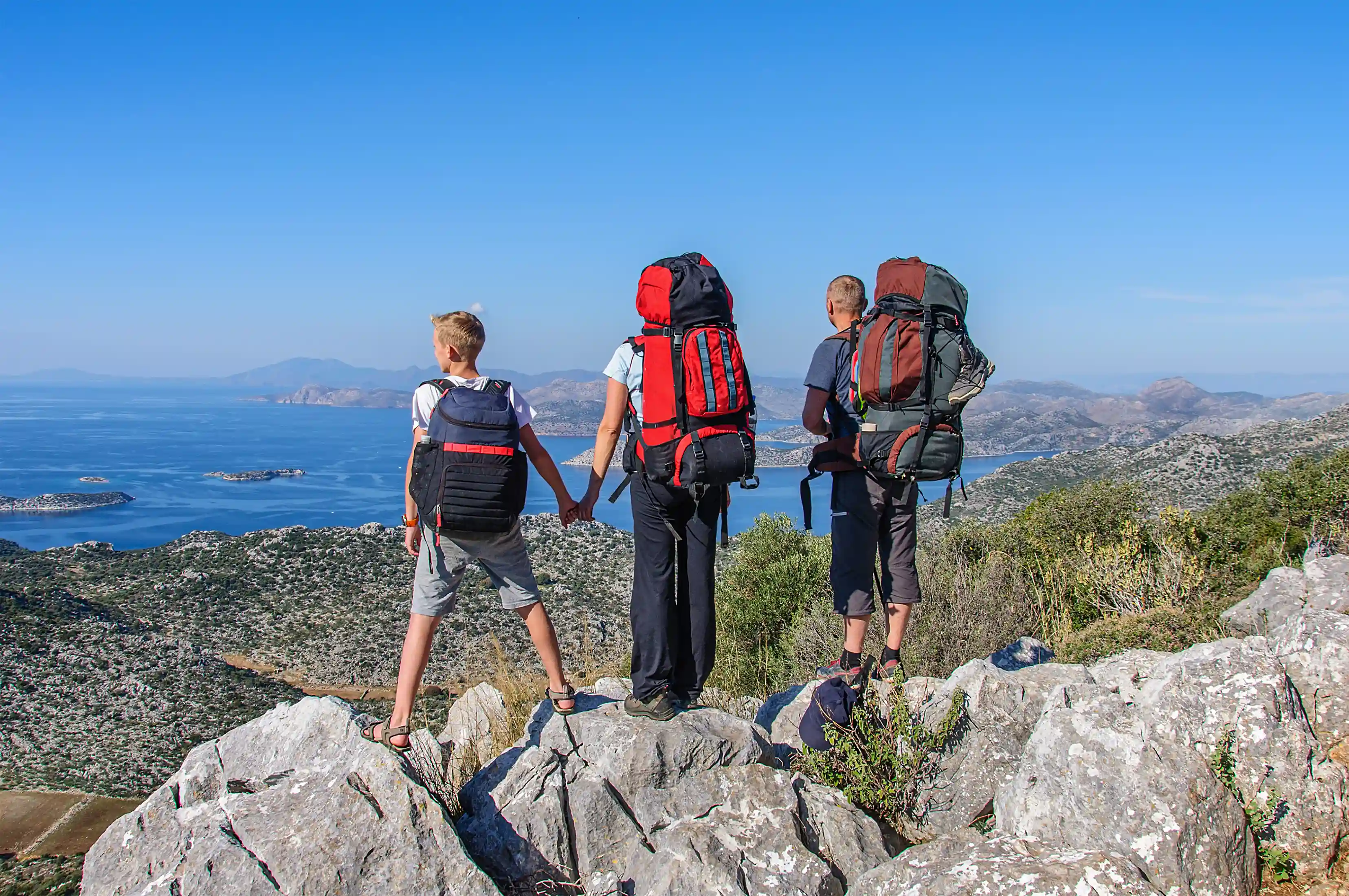 Three hikers with large backpacks standing on rocky terrain overlooking a coastal landscape with blue water and islands.