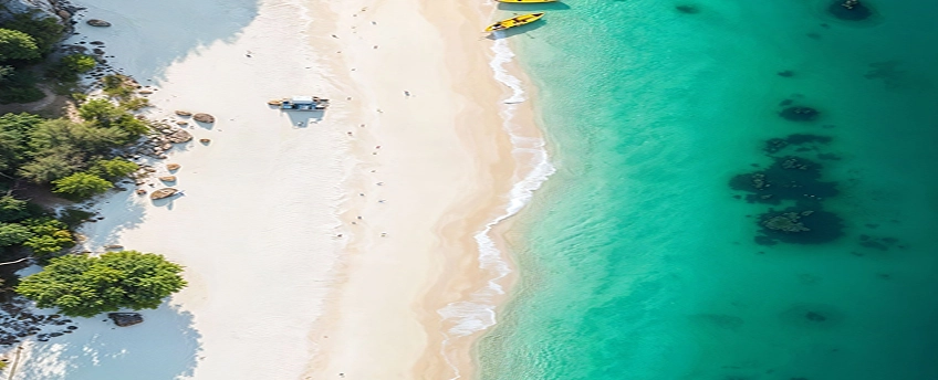 Aerial view of a sandy beach with turquoise water, a yellow kayak near the shore, and green trees lining the beach.