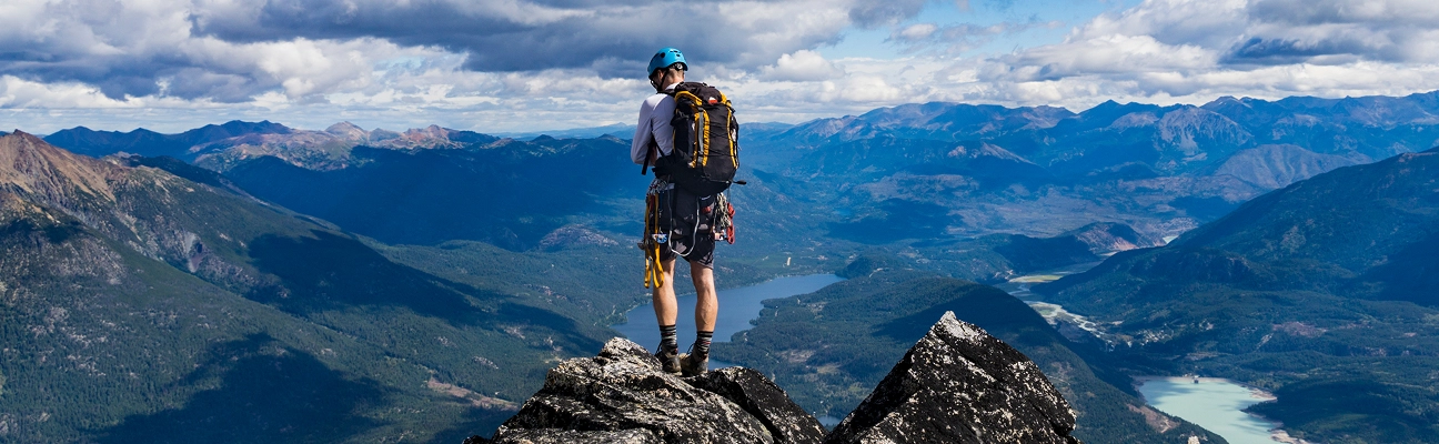 Hiker wearing a blue helmet and yellow-accented backpack standing on a rocky peak overlooking a vast mountainous landscape with lakes under a partly cloudy sky.