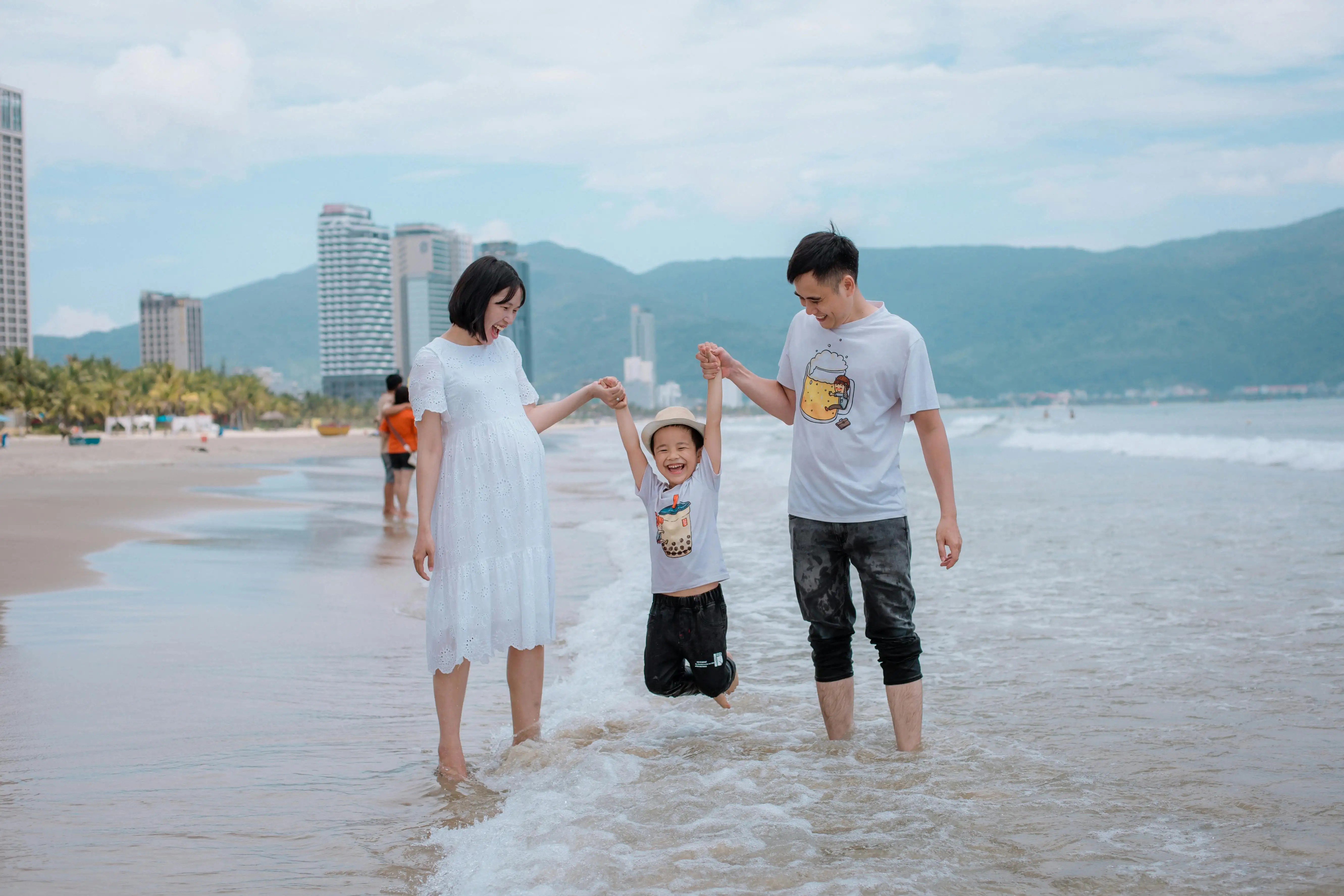 Smiling parents holding their child's hands while the child jumps at the beach shore with city buildings and mountains in the background.